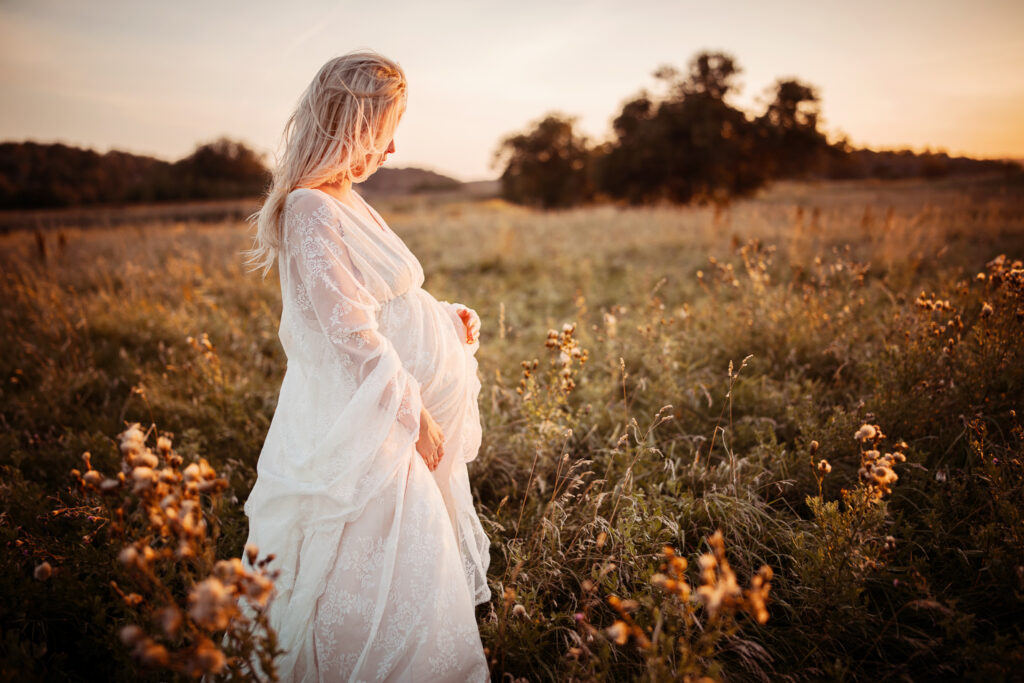 Gravid kvinna i vit klänning i blomstrande äng vid solnedgång.