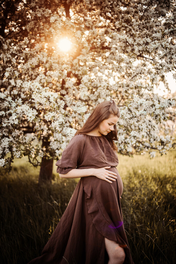 Gravid kvinna i brunt vid blommande träd i motljus.